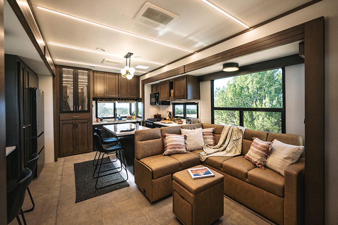 Modern Bighorn RV interior with a cozy brown sectional sofa, pillows, and blanket, a kitchen area with dark cabinets, bar stools, and large windows showcasing green trees outside for a bright, inviting atmosphere.