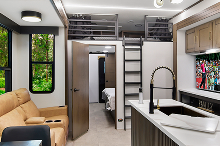 Modern RV interior with beige recliners, a kitchen island, appliances, overhead storage, and large windows showing a green forest. A TV above the stove displays a sports crowd. Some bunkhouses offer similar layouts with a bedroom visible through an open door.