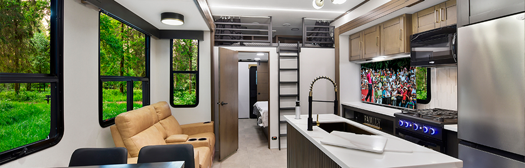 Modern RV interior with beige recliners, a kitchen island, appliances, overhead storage, and large windows showing a green forest. A TV above the stove displays a sports crowd. Some bunkhouses offer similar layouts with a bedroom visible through an open door.