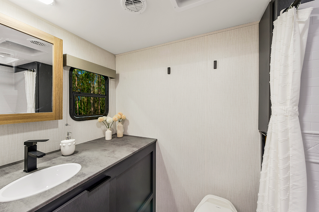 A modern bathroom in a travel trailer features a sink, black faucet, countertop, toilet, white shower curtain, wall hooks, and a window with a nature view. Light walls and minimalist decor create an innovative, clean, and bright space.