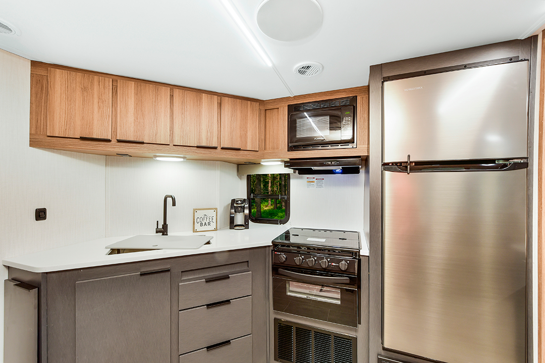 Modern RV kitchen with innovative wood cabinets, a stainless steel refrigerator, microwave, stove, oven, sink, coffee maker, and a small sign reading Coffee Bar on the counter under bright lighting—perfect for any travel trailer adventure.