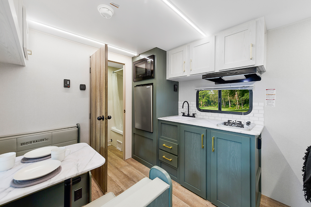 Modern RV interior featuring a compact dining area, a kitchenette with green cabinets, and full amenities—perfect for couples coaches—with a view of greenery through the window; a bathroom is visible through an open door.
