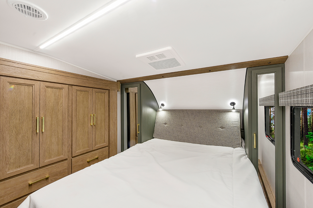 A cozy RV bedroom in one of our couples coaches features a neatly made bed, wooden cabinets with gold handles on the left, modern reading lights above the headboard, and a window showing greenery on the right.