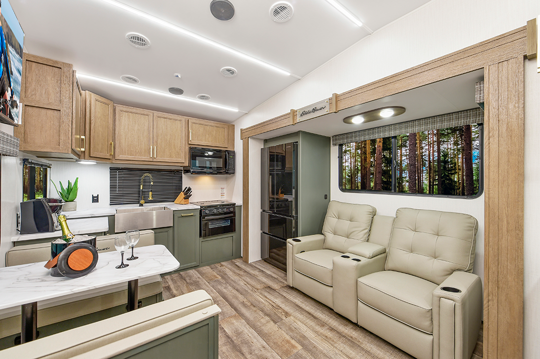 Modern couples coach interior with wood and sage green cabinetry, a marble-look table set for wine, two beige recliners, a kitchen with sink and stove, and large windows overlooking the forest. The space is well-lit and cozy.