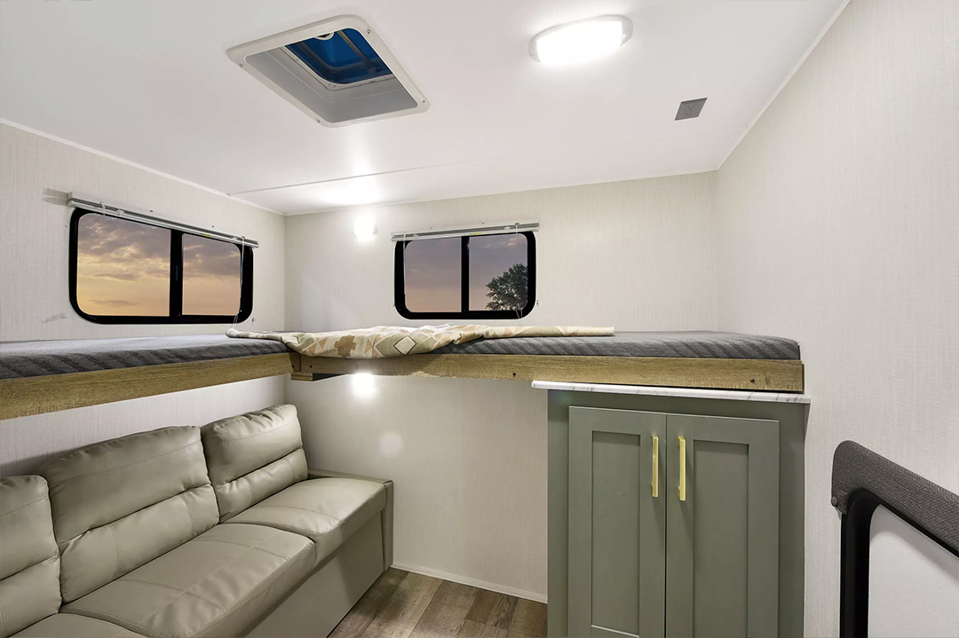 Modern RV interior with light-colored walls, a bunk bed above a beige sofa, two windows showing a sunset, and a green cabinet with gold handles on wood flooring. Ceiling lights, a small skylight, and classic bunkhouses touches complete the space.