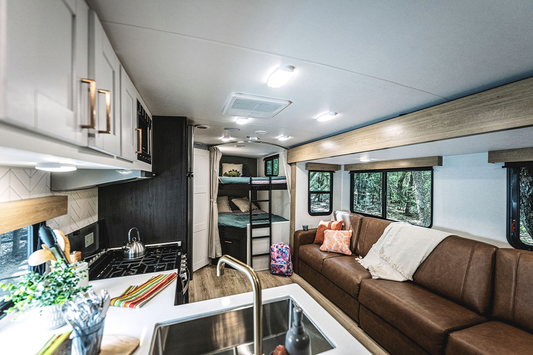 Interior of a modern RV bunkhouse, featuring a brown leather couch with throw pillows and a blanket, a sink and stove in the foreground, and cozy bunk beds with curtains in the back. Large windows reveal a scenic wooded landscape.