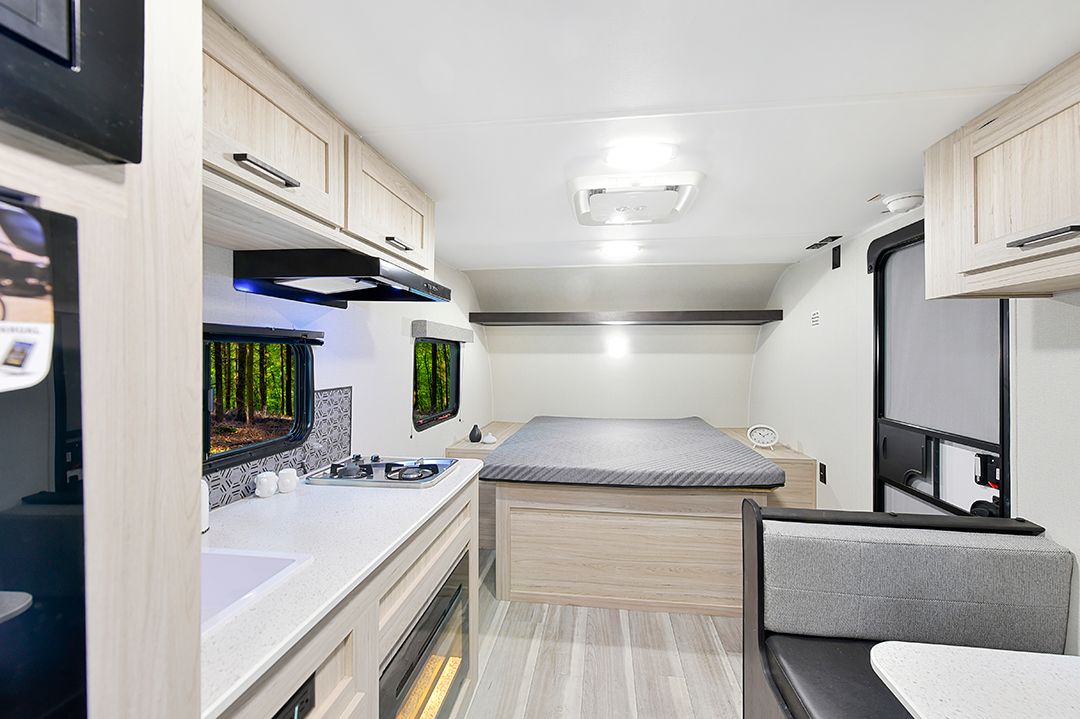 Interior of an innovative travel trailer, featuring a compact kitchen with stove and sink, a bed with gray bedding, wooden cabinets, a window framing a forest view, and a cushioned bench seat.