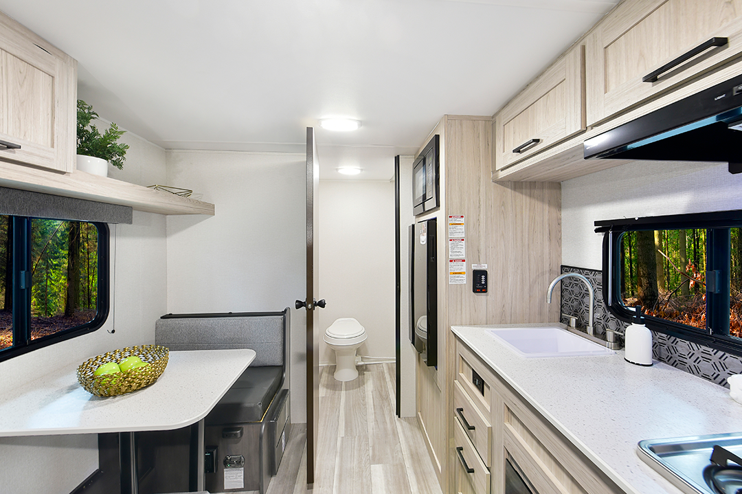 Modern RV interior featuring a kitchen with a sink, stovetop, cabinets, and countertop, a dining area with table and bowl of fruit—ideal for couples coaches—and a view into a small bathroom. Windows reveal a forest outside.