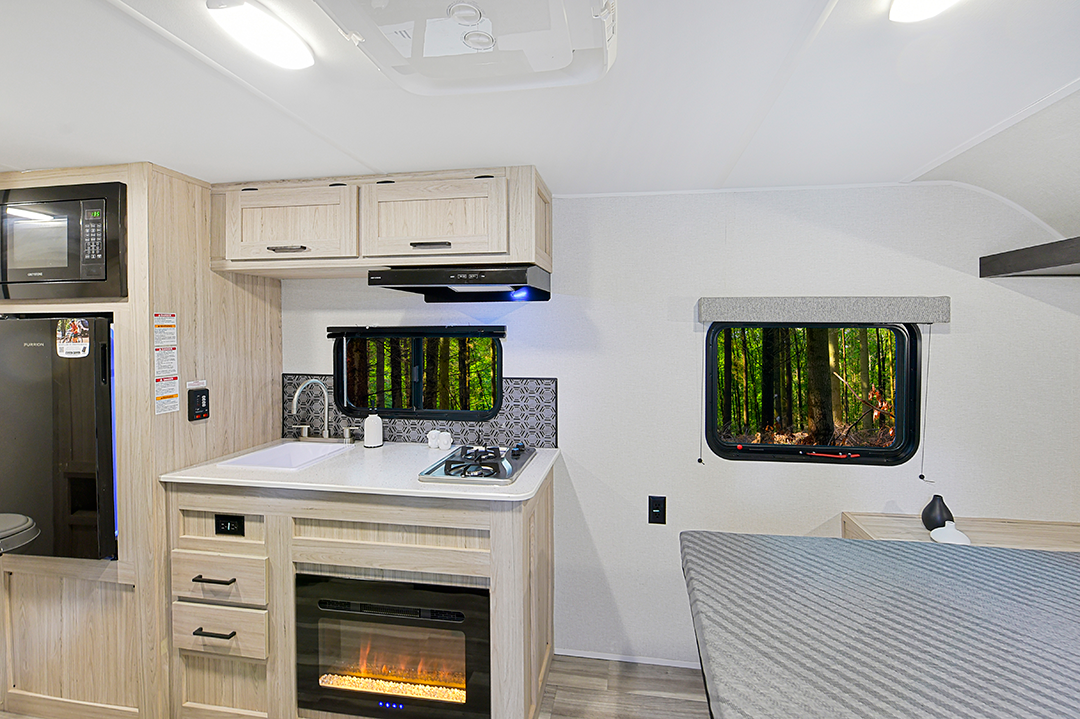 Modern RV interior with light wood cabinets, a kitchen area including a sink, stovetop, and electric fireplace. Perfect for couples coaches, a cozy bed sits on the right, while windows reveal a view of the green forest outside.