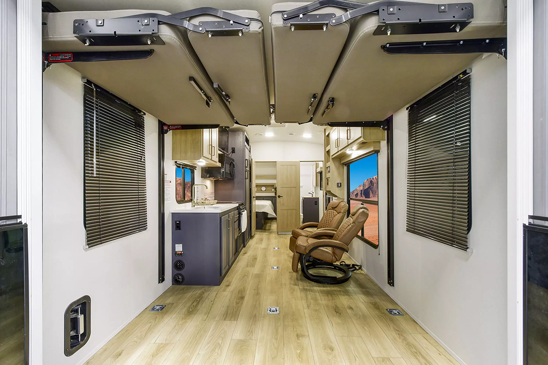 Interior of a modern RV with wood flooring, a kitchenette on the left, two brown recliners facing a large window with a scenic desert view, and fold-down beds above—ideal for couples coaches seeking comfort and adventure.