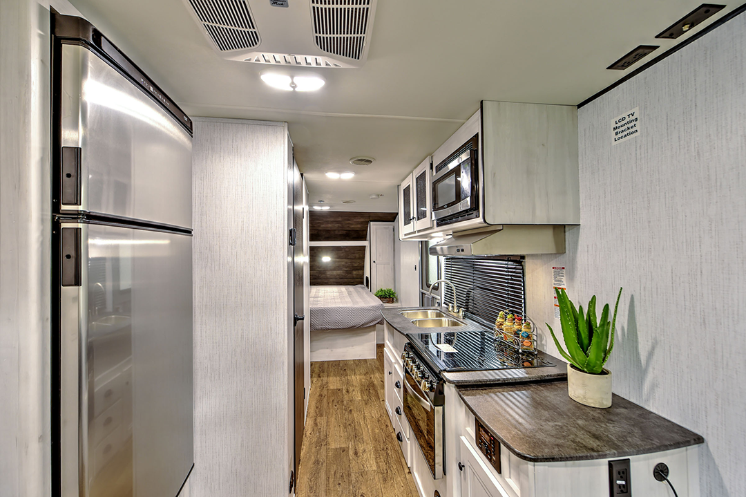 A modern RV kitchen in one of our couples coaches features stainless steel appliances, double sink, stove, microwave, and a countertop with a potted plant. Light wood cabinets and flooring lead to a cozy bed in the rear.