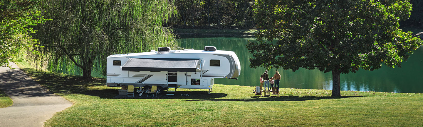 A family stands near a white fifth-wheel camper parked on a grassy lakeshore.
